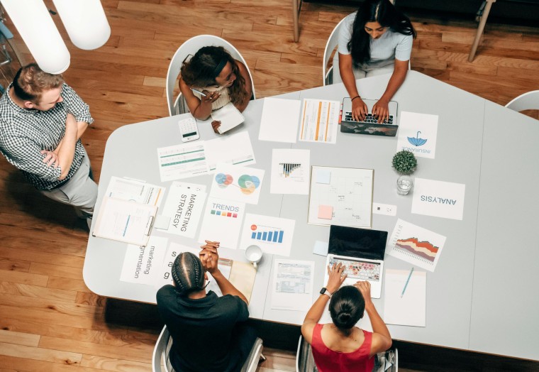 bird's eye view of 5 people sitting around a meeting room table with paper and computers on it