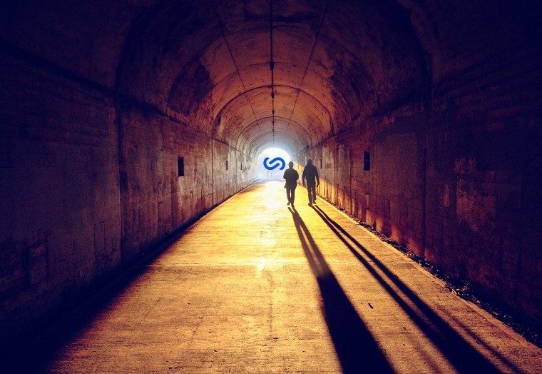 view from behind two people walking in a dark tunnel with a bright silverstripe logo at the end of the tunnel